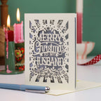 A laser-cut Christmas card with the text 'Merry Christmas To My Husband' in an intricate snowflake and floral design, displayed on a table with candles in the background.