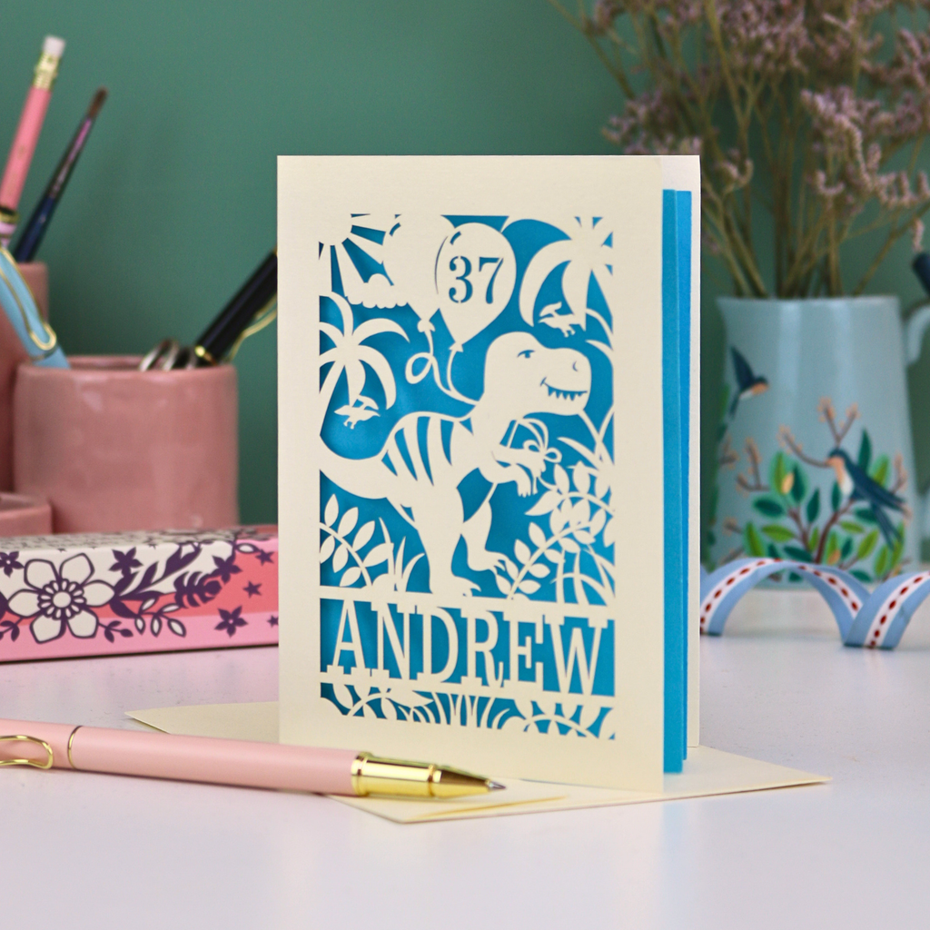 Personalised male birthday Card with dinosaur design and 'Andrew' and '37' text on a desk with stationery items.