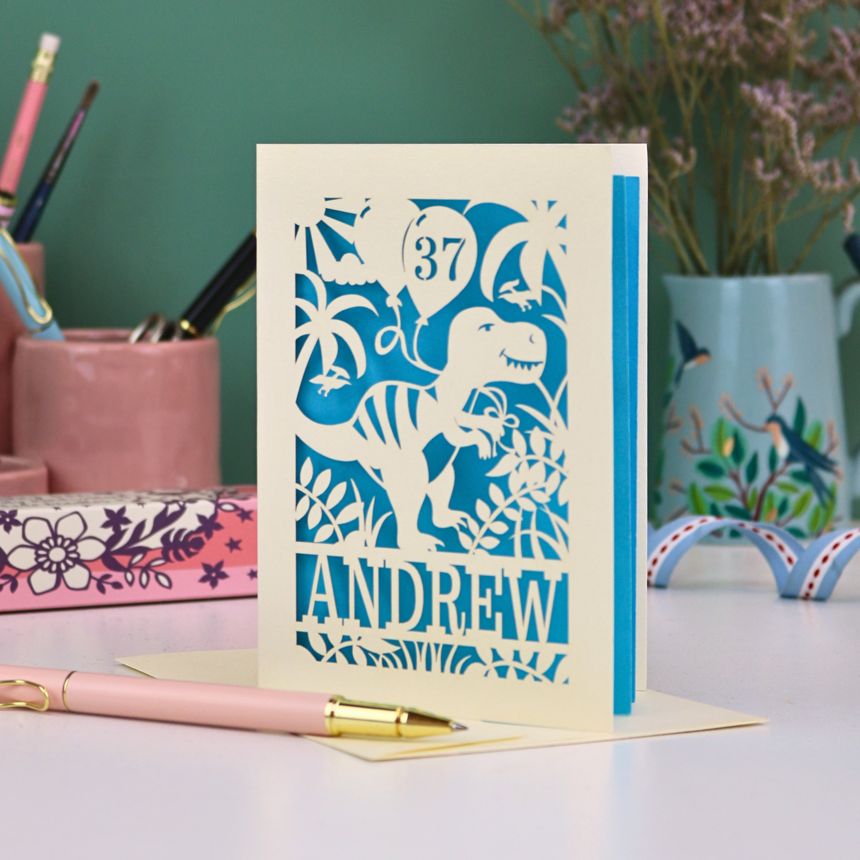 Personalised male birthday Card with dinosaur design and 'Andrew' and '37' text on a desk with stationery items.