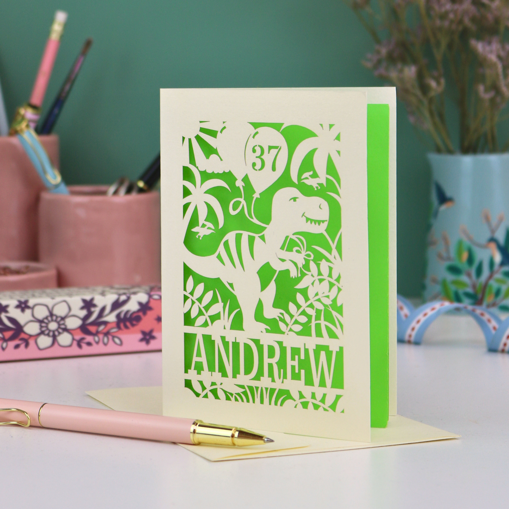 Personalised husband birthday Card with dinosaur design and 'Andrew' and '37' text on a desk with stationery items.