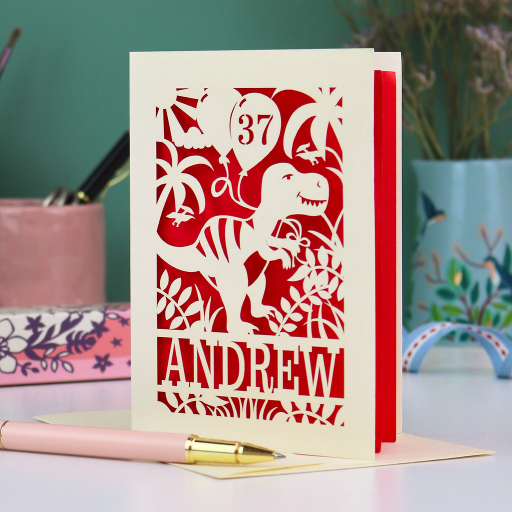 Personalised male birthday Card with dinosaur design and 'Andrew' and '37' text on a desk with stationery items.