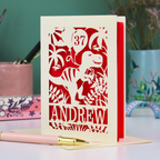 Personalised male birthday Card with dinosaur design and 'Andrew' and '37' text on a desk with stationery items.
