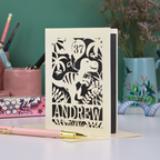 Personalised masculine birthday Card with dinosaur design and 'Andrew' and '37' text on a desk with stationery items.
