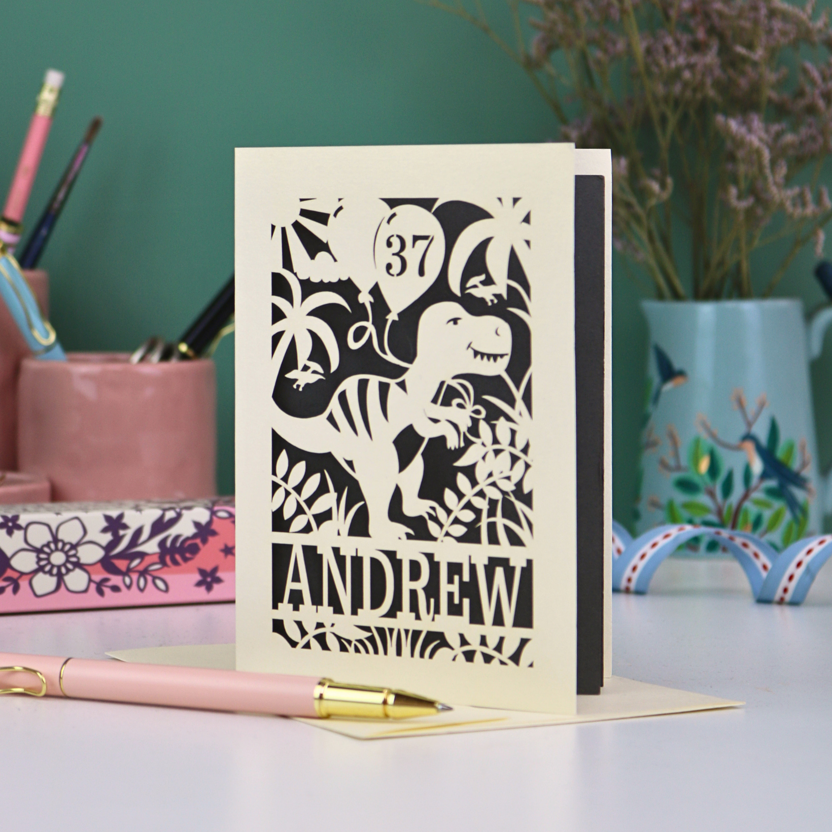 Personalised masculine birthday Card with dinosaur design and 'Andrew' and '37' text on a desk with stationery items.