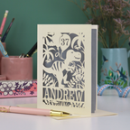 Personalised male birthday Card with laser cut dinosaur design and 'Andrew' and '37' text on a desk with stationery items.