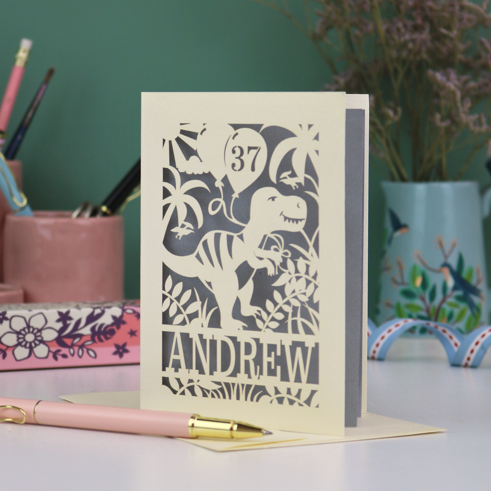 Personalised male birthday Card with laser cut dinosaur design and 'Andrew' and '37' text on a desk with stationery items.