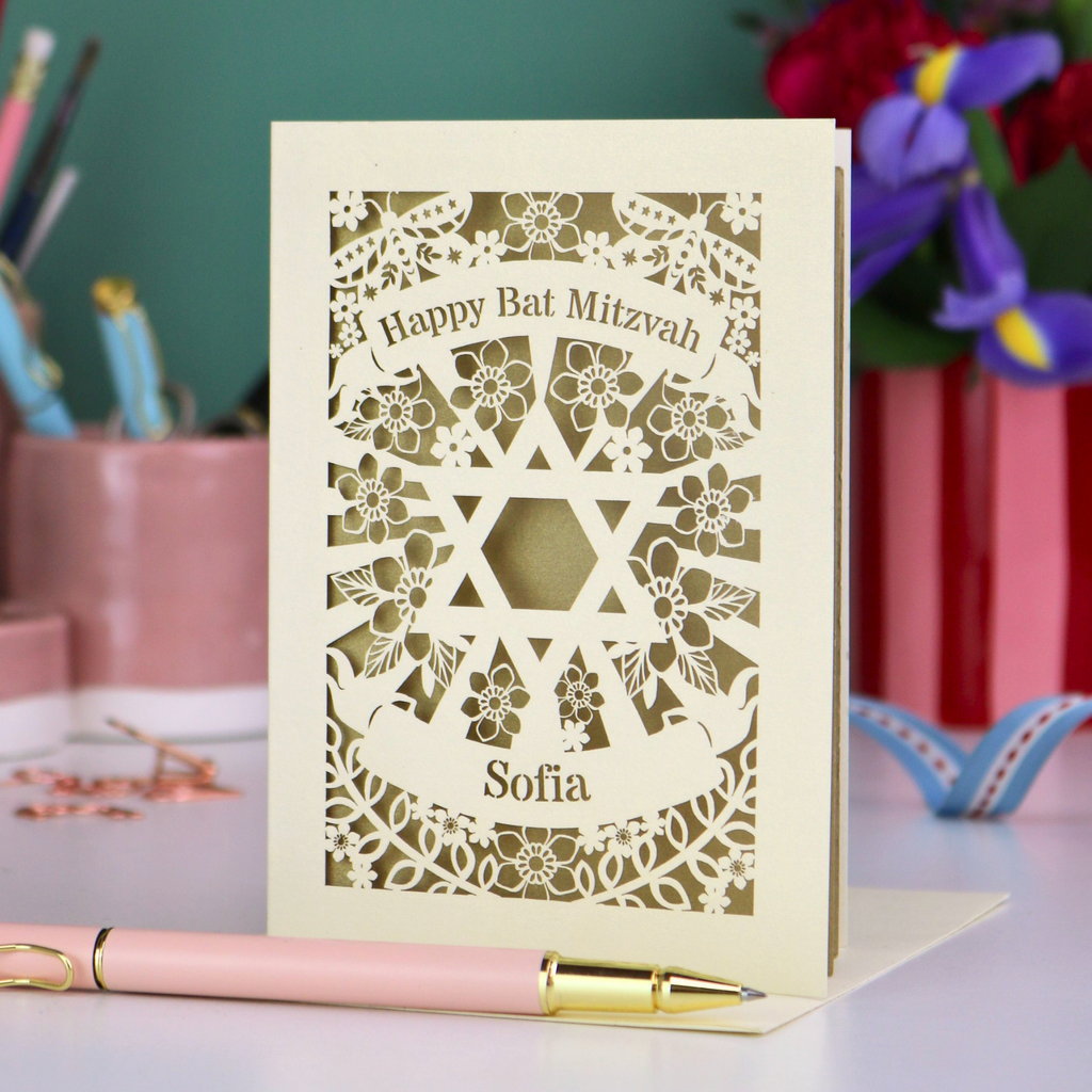 Laser-cut 'Happy Bat Mitzvah' card with decorative patterns on a table with stationery items.