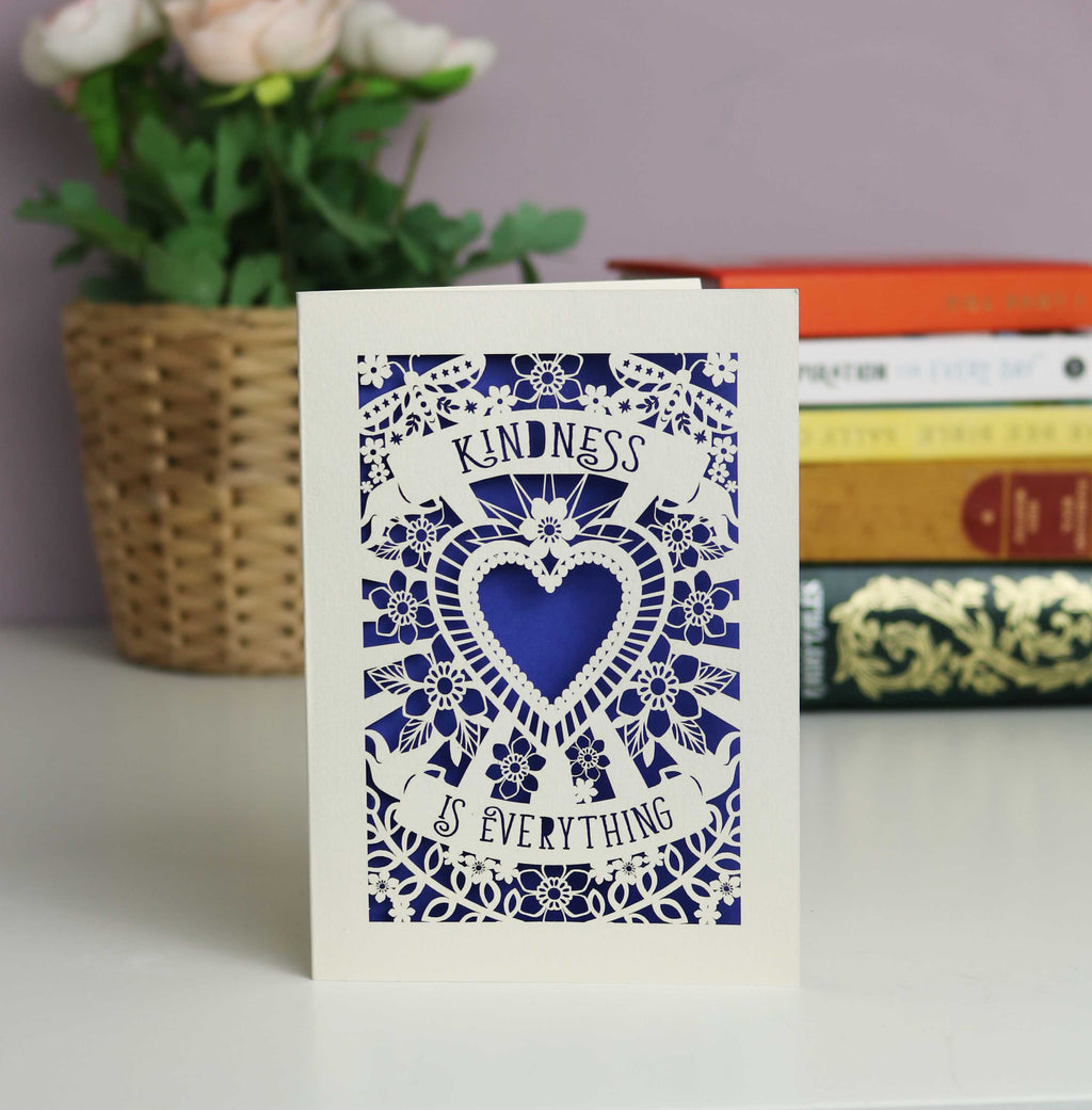 Elegant papercut card in cream with a dark blue background. Text in 2 banners says "Kindness is everything" surrounding a central heart with flowers and butterflies.