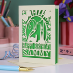Personalized 21st birthday card with number 21 and decorative elements, and text "Happy Birthday Sammy" on a desk with stationery items.