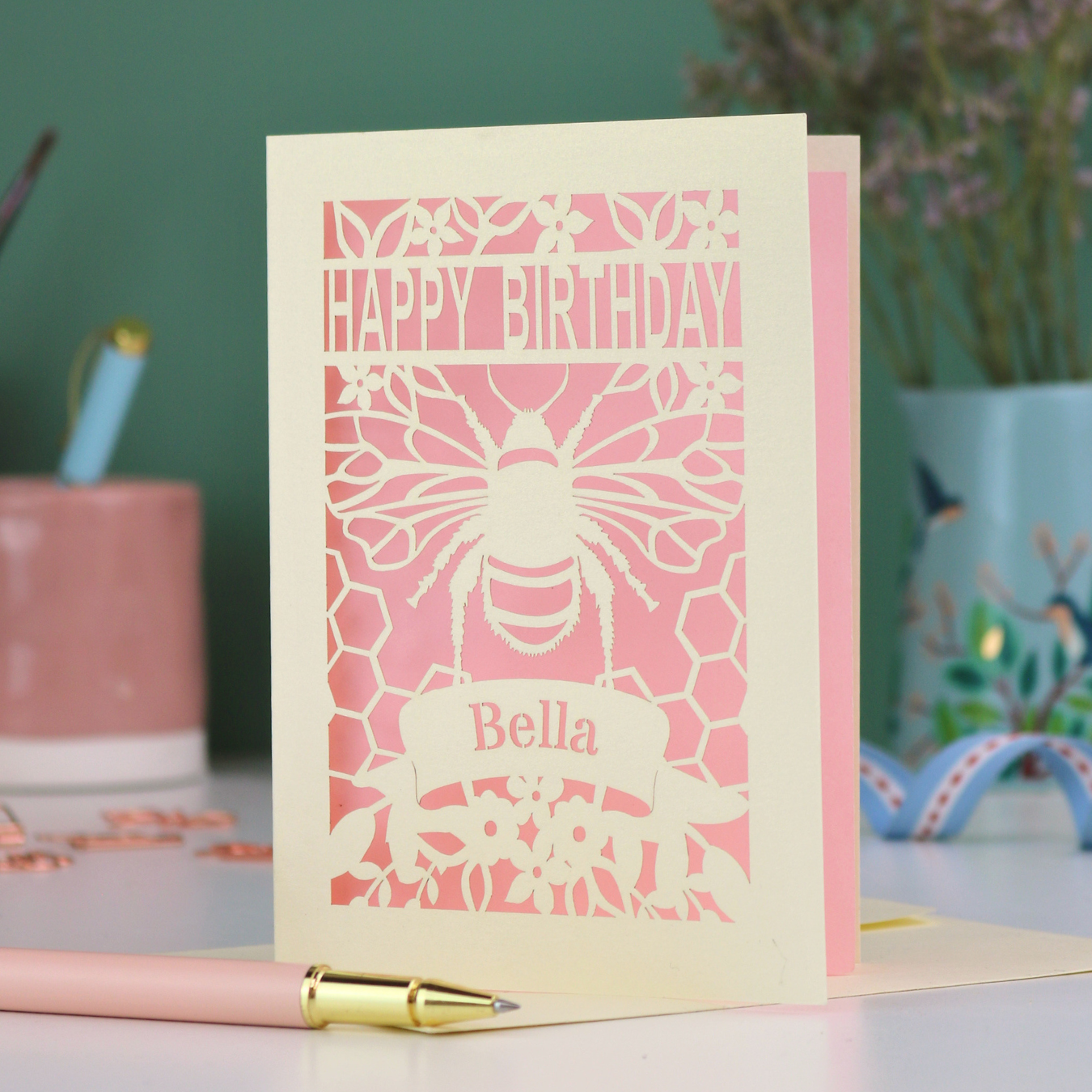 A laser cut birthday card with cut out bee and honeycomb design and text "Happy Birthday", personalised with the name "Bella" in a stencil font on a banner.