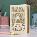 A laser cut birthday card with cut out bee and honeycomb design and text "Happy Birthday", personalised with the name "Bella" in a stencil font on a banner.