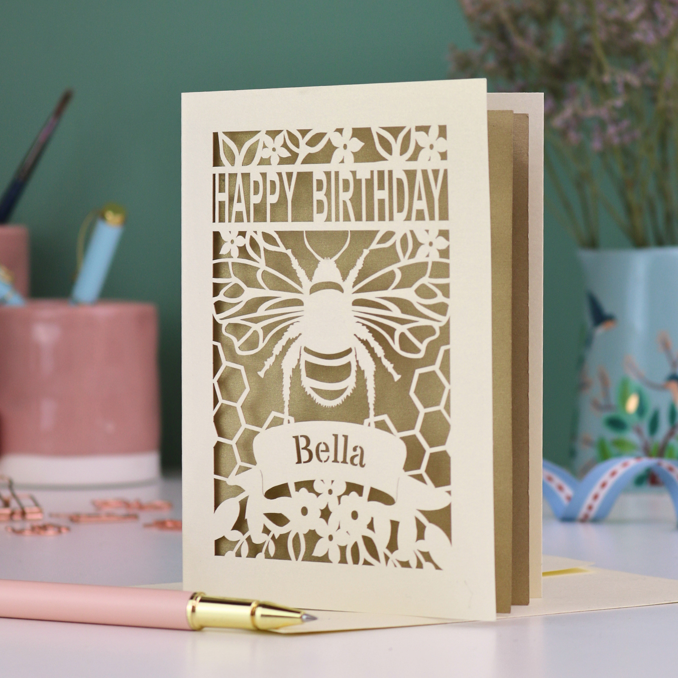 A laser cut birthday card with cut out bee and honeycomb design and text "Happy Birthday", personalised with the name "Bella" in a stencil font on a banner.