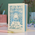 A laser cut birthday card with cut out bee and honeycomb design and text "Happy Birthday", personalised with the name "Bella" in a stencil font on a banner.