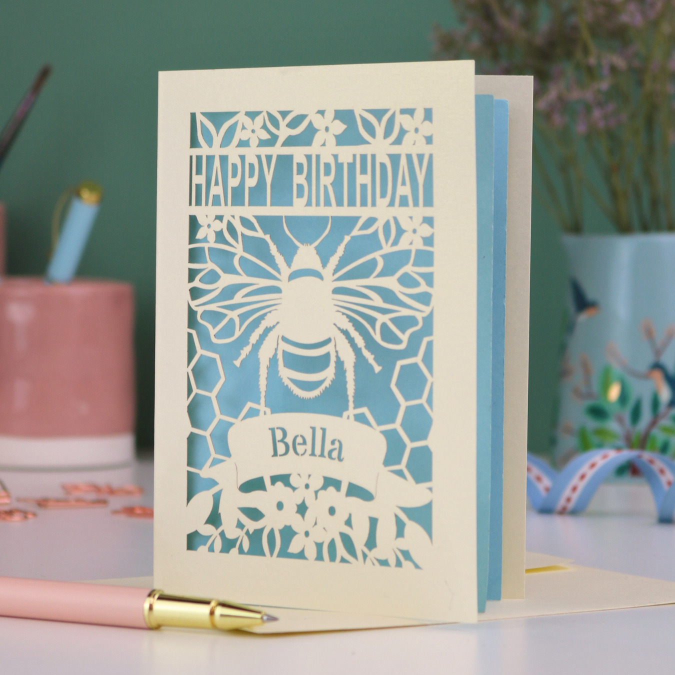 A laser cut birthday card with cut out bee and honeycomb design and text "Happy Birthday", personalised with the name "Bella" in a stencil font on a banner.