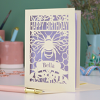 A laser cut birthday card with cut out bee and honeycomb design and text "Happy Birthday", personalised with the name "Bella" in a stencil font on a banner.