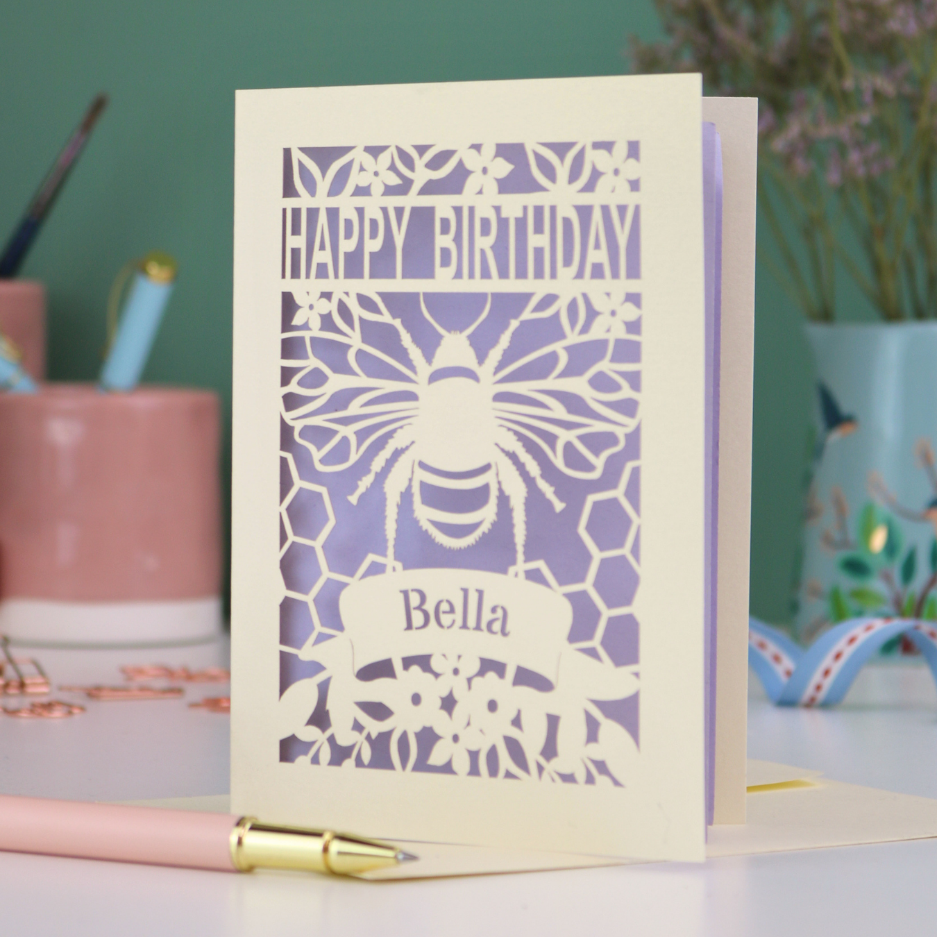 A laser cut birthday card with cut out bee and honeycomb design and text "Happy Birthday", personalised with the name "Bella" in a stencil font on a banner.