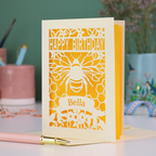 A laser cut birthday card with cut out bee and honeycomb design and text "Happy Birthday", personalised with the name "Bella" in a stencil font on a banner.