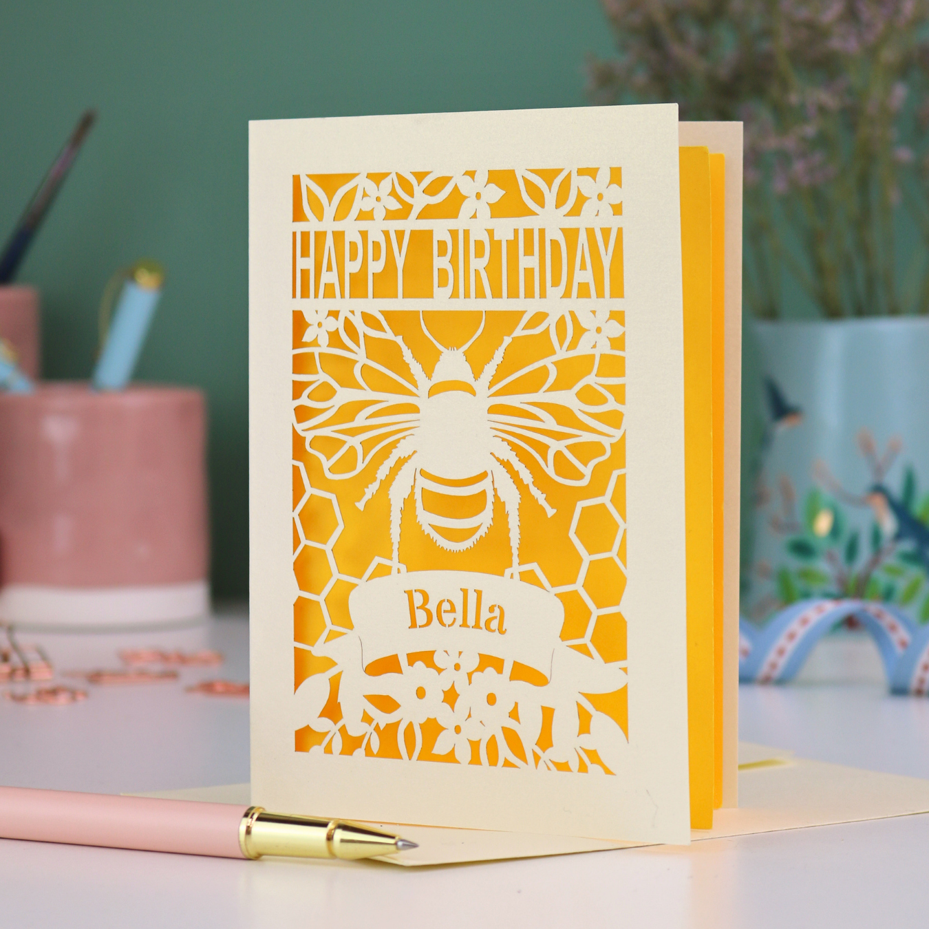 A laser cut birthday card with cut out bee and honeycomb design and text "Happy Birthday", personalised with the name "Bella" in a stencil font on a banner.