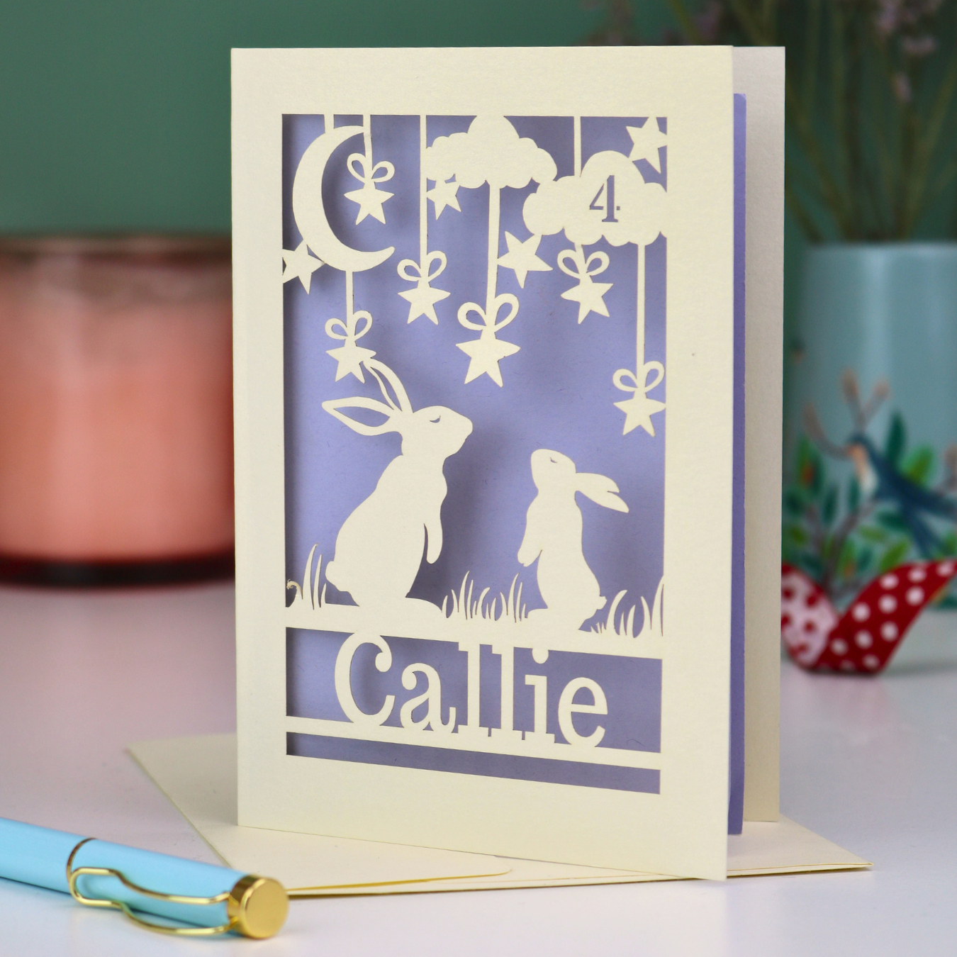Laser cut birthday card with cut-out rabbits looking up at the moon design and 'Callie' text on a lilac background