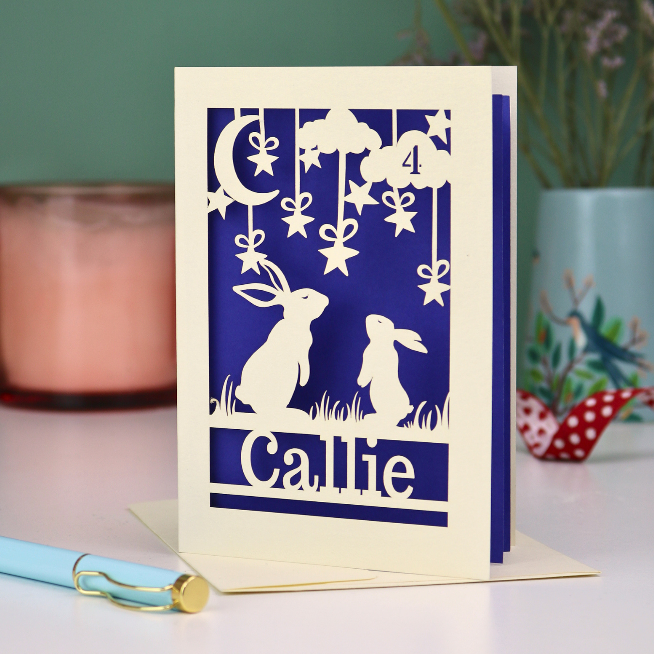 Paper cut birthday card with cut-out rabbits looking up at the moon design and 'Callie' text on a violet background
