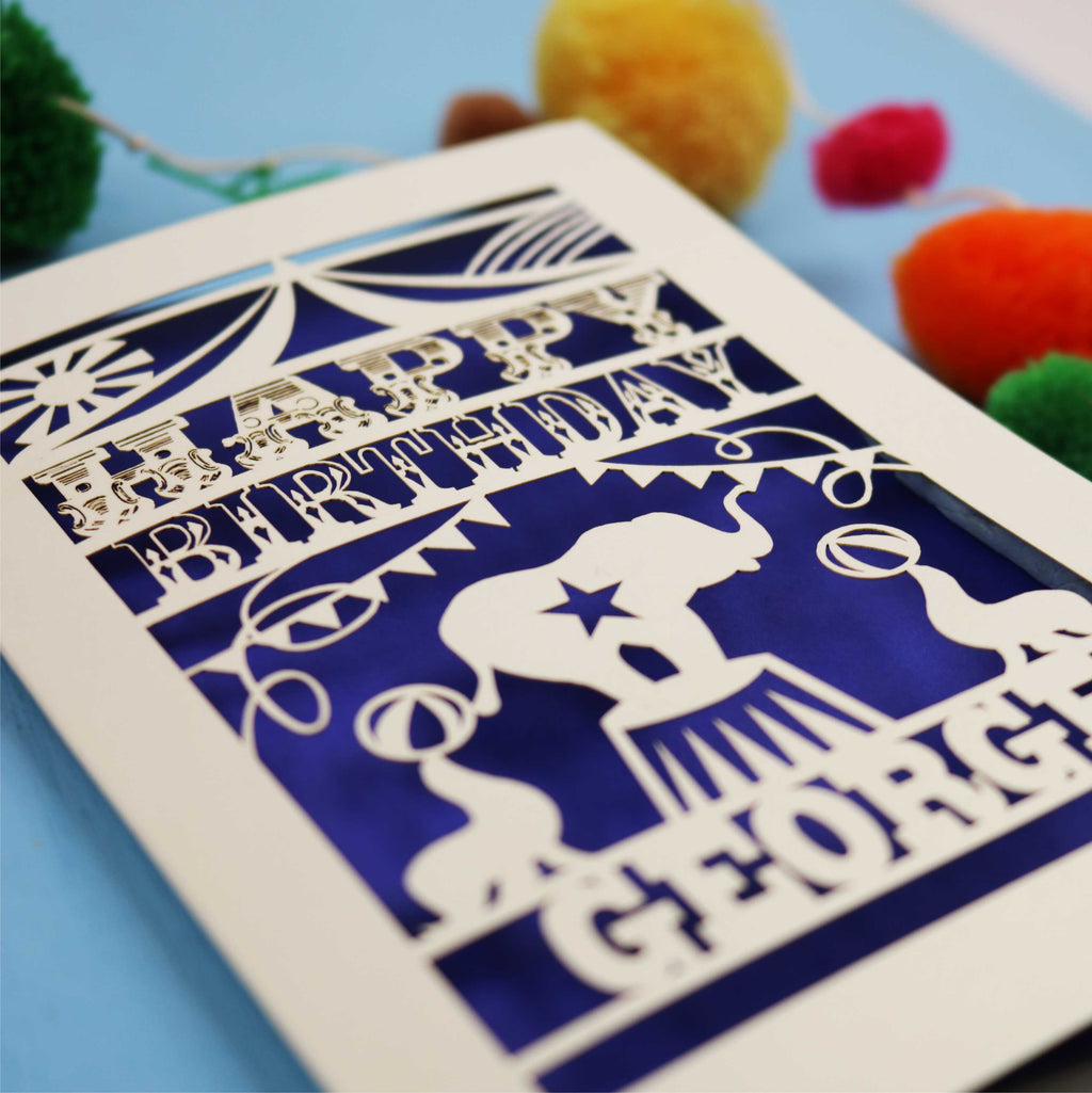 Personalised laser cut Circus Birthday card with 'Happy Birthday George' text on a blue background with colourful pom-poms.