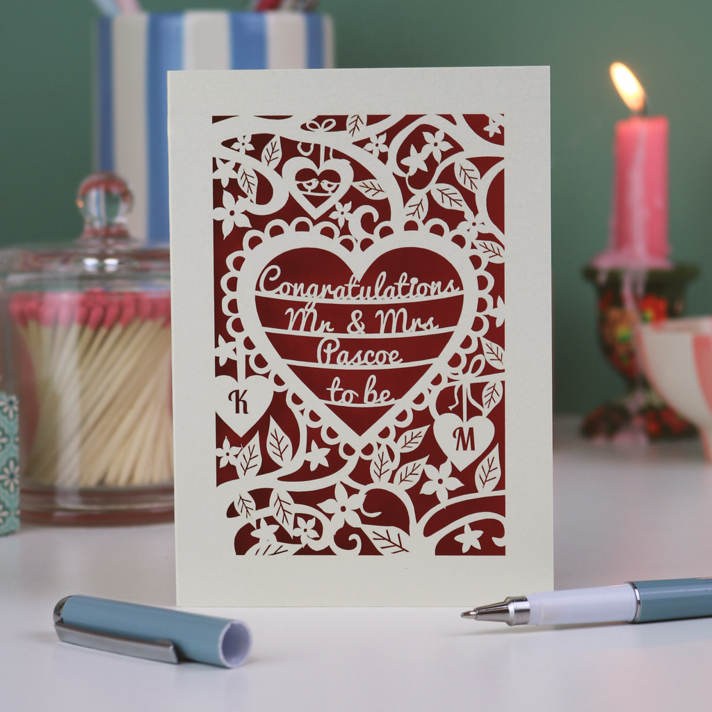A laser cut engagement card with floral heart design, sitting on a white desk with ribbons, pen and pots on a green background