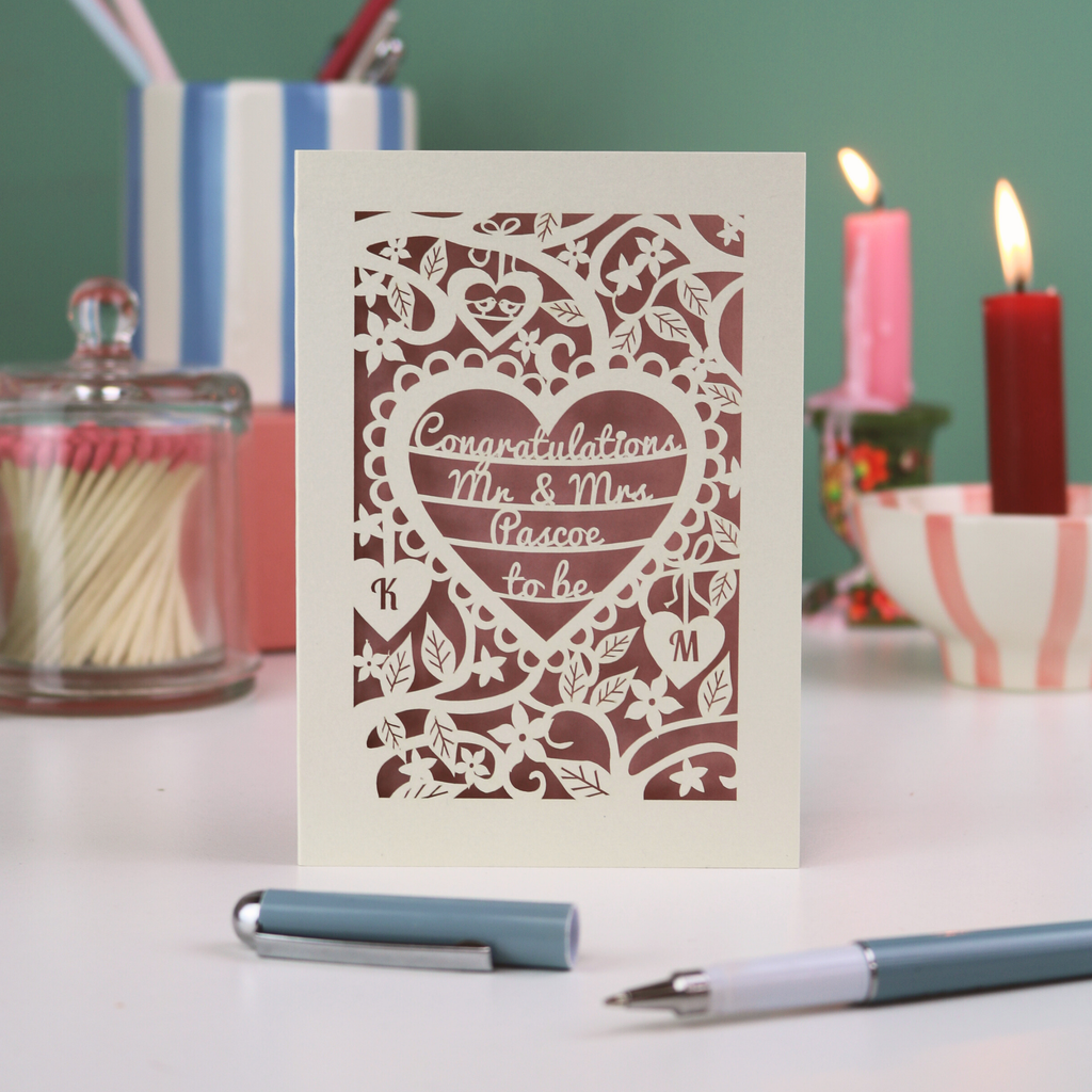 A laser cut engagement card with floral heart design, sitting on a white desk with candles, pen and pots on a green background