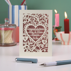 A laser cut engagement card with floral heart design, sitting on a white desk with candles, pen and pots on a green background