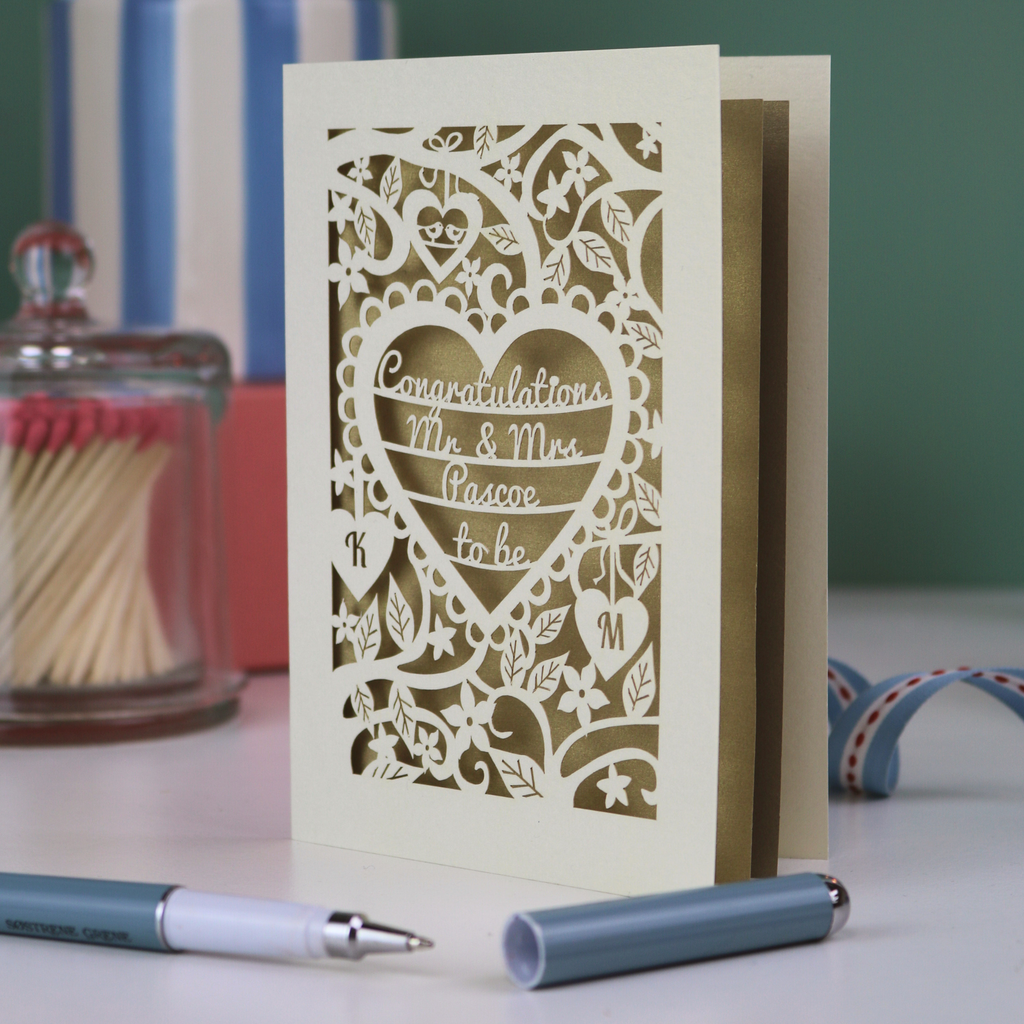 A laser cut engagement card with floral heart design, sitting on a white desk with ribbons, pen and pots on a green background