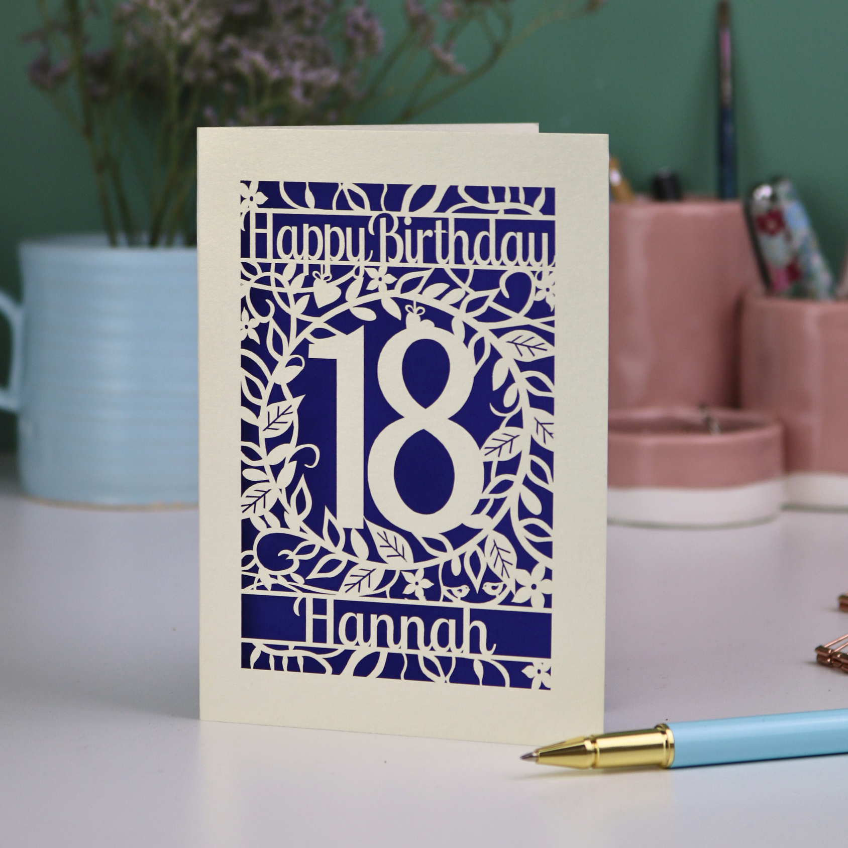 Cut out Birthday card with 'Happy Birthday 18 Hannah' on a table with a pen and decorative items.