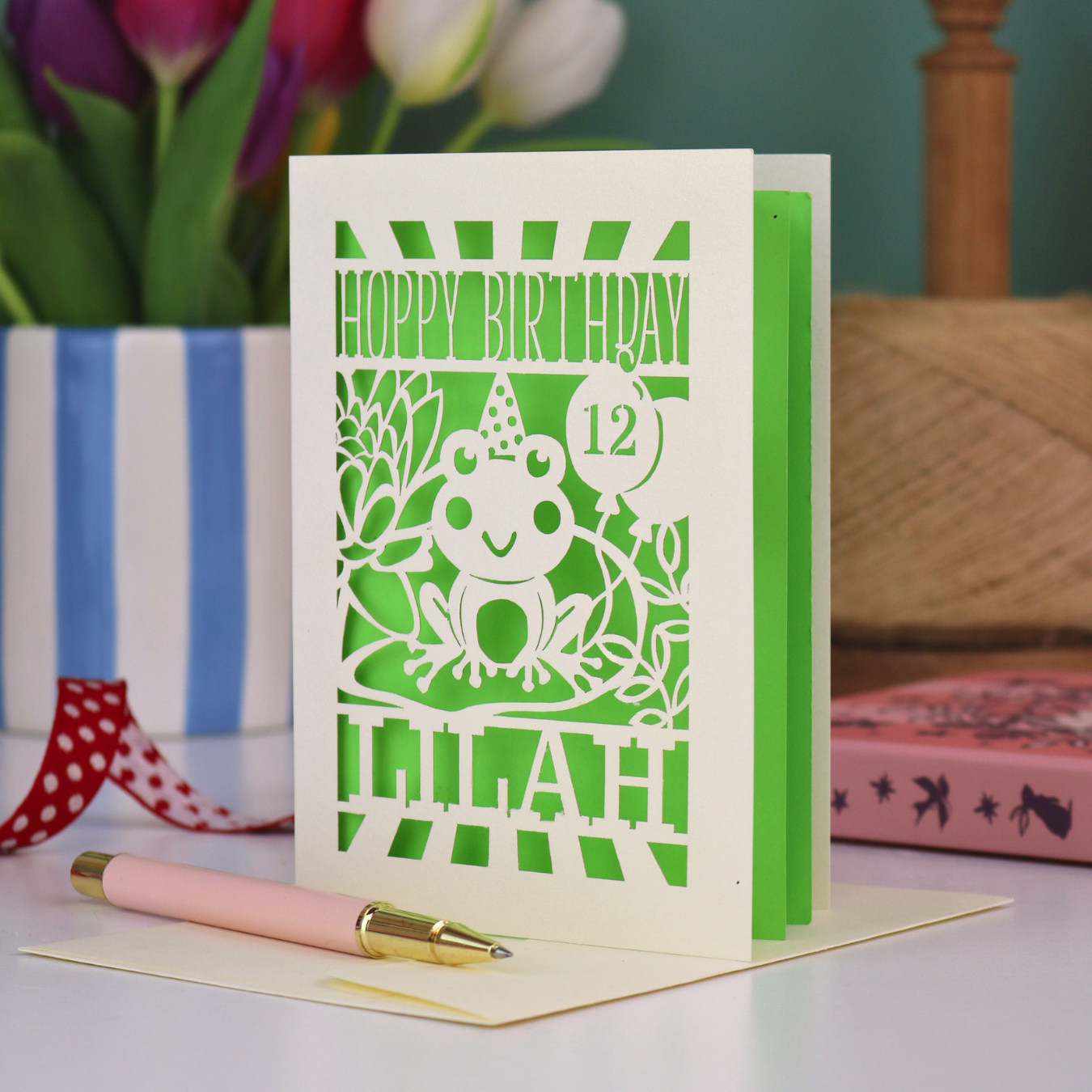 A childrens birthday card, personalised with a name and age, featuring a laser cut frog design and the text "Hoppy Birthday"