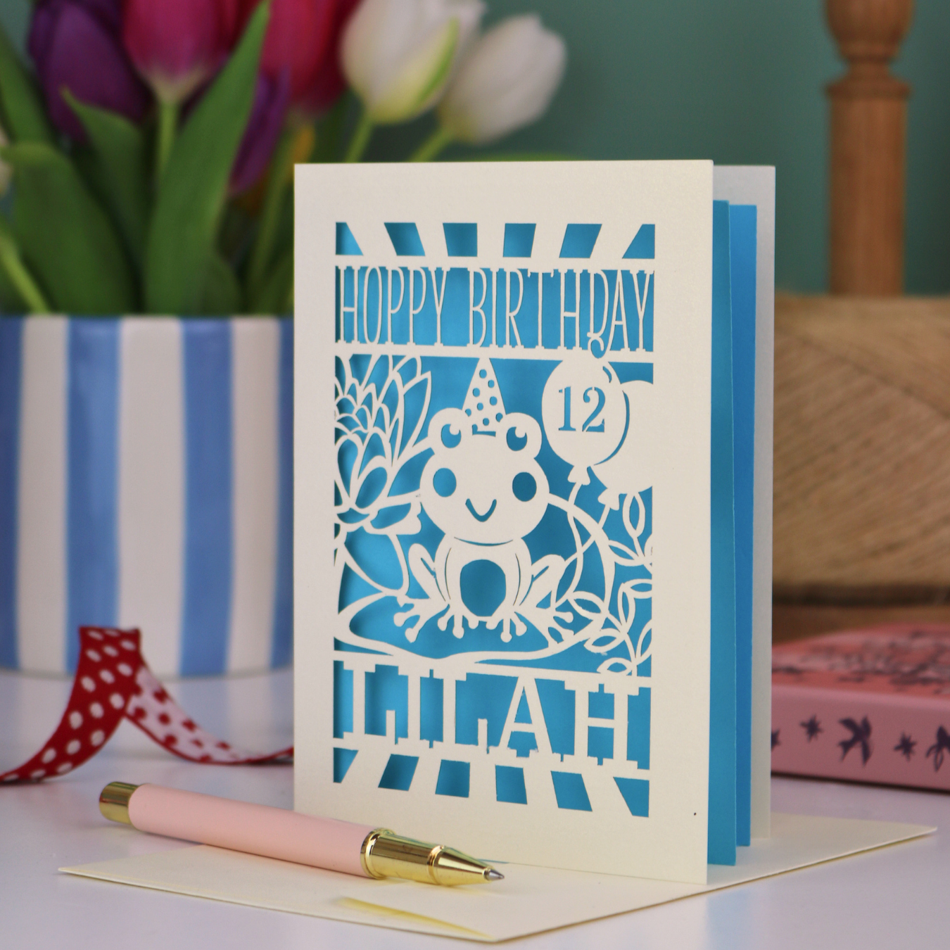A child's birthday card, personalised with a name and age, featuring a laser cut frog design and the text "Hoppy Birthday"