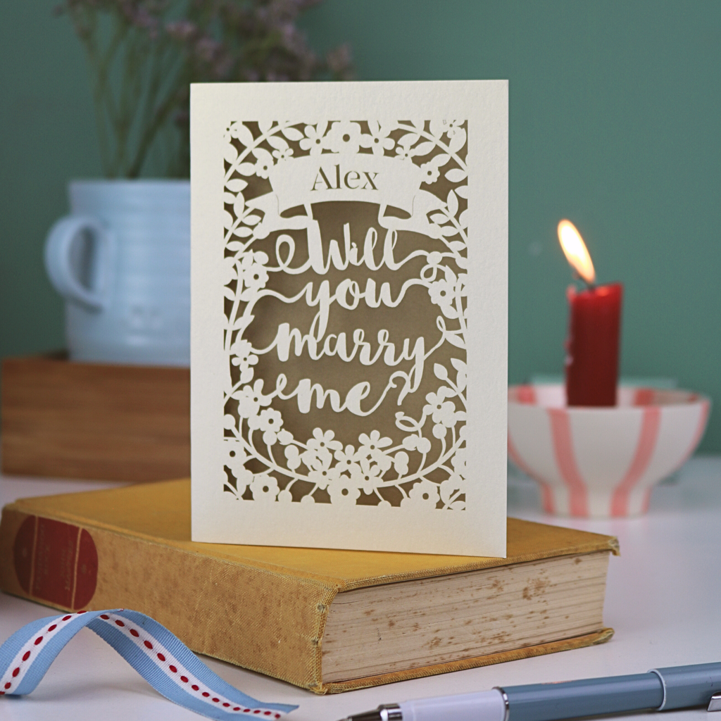 Proposal card with 'Will you marry me?' on a book with a candle and cup in the background