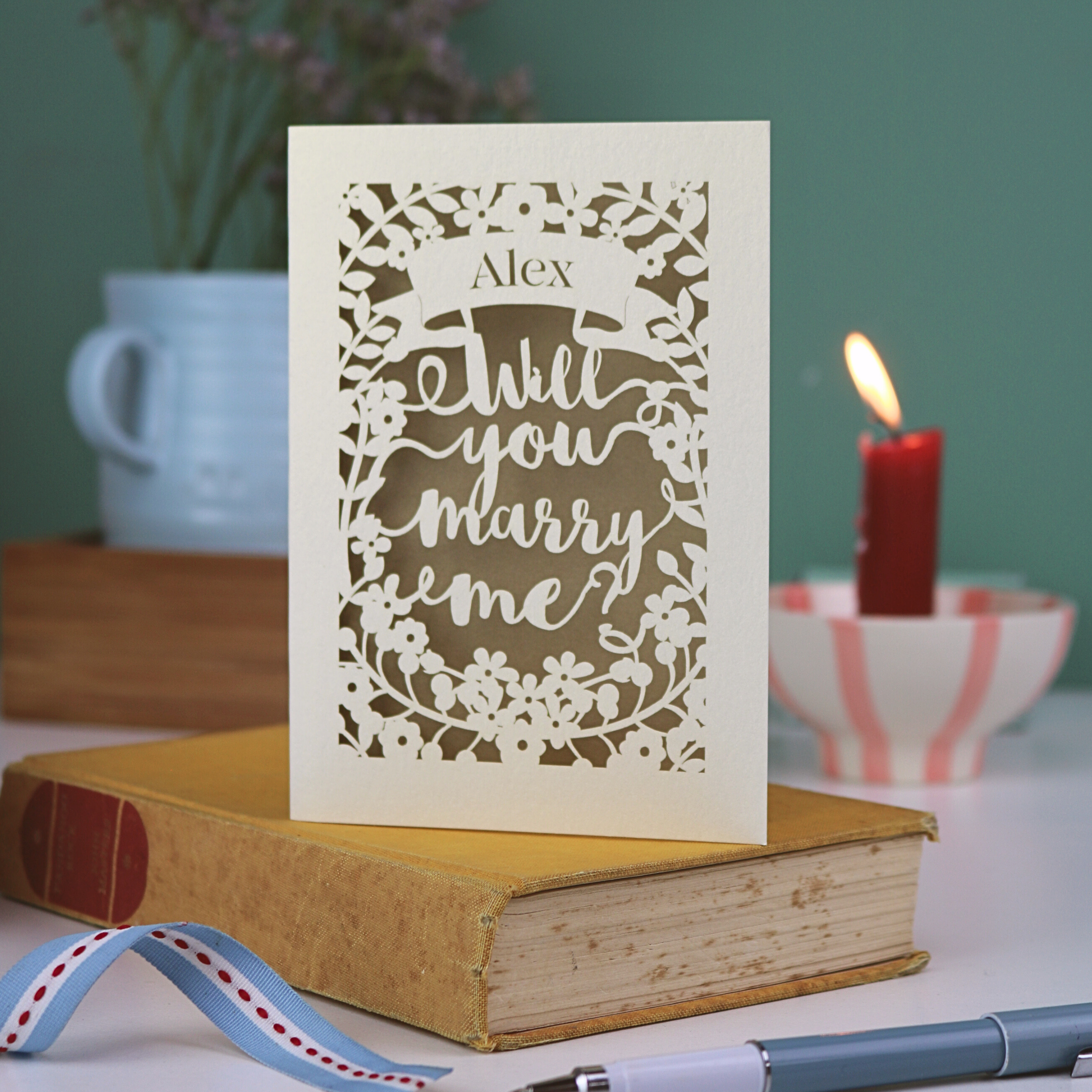 Proposal card with 'Will you marry me?' on a book with a candle and cup in the background