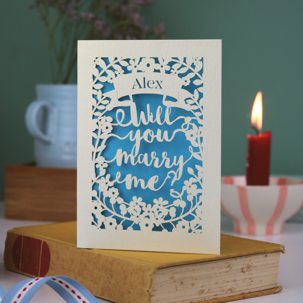 Proposal card with 'Will you marry me?' on a book with a candle and flowers in the background