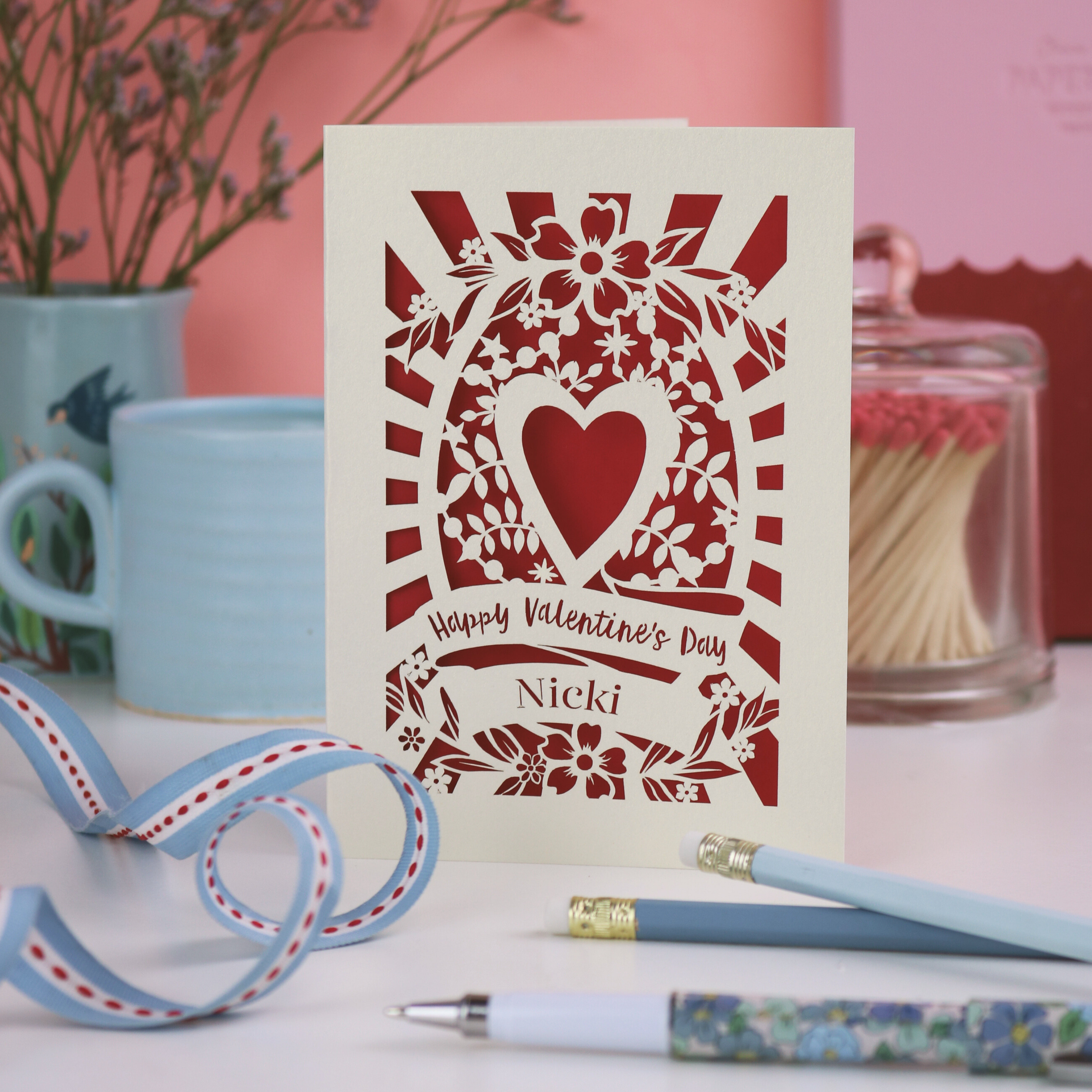 A personalised laser cut Valentine's Day card with decorative heart design on a table with stationery items.