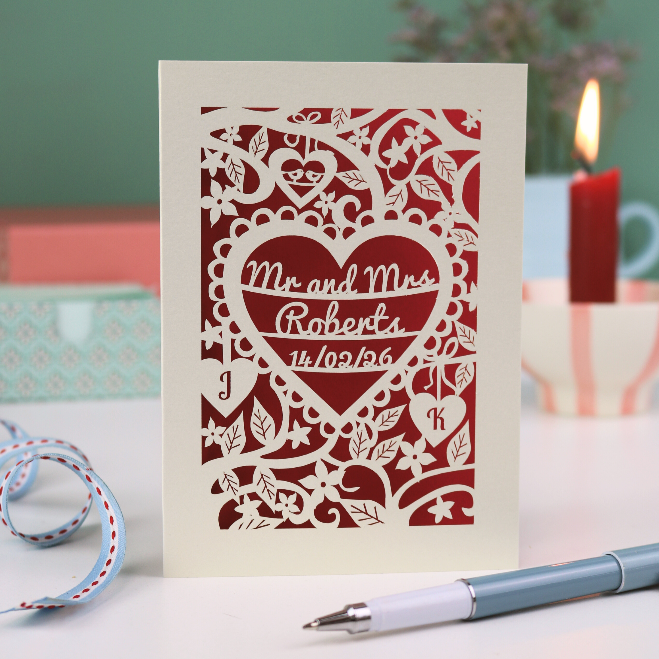 A personalised wedding card with floral heart design, sitting on a white desk with ribbons, stationery items and plant on a green background. Card is cream with "Mr and Mrs Roberts, 14/02/26" on 3 lines of text inside a heart shape with intricate floral cut out border.
