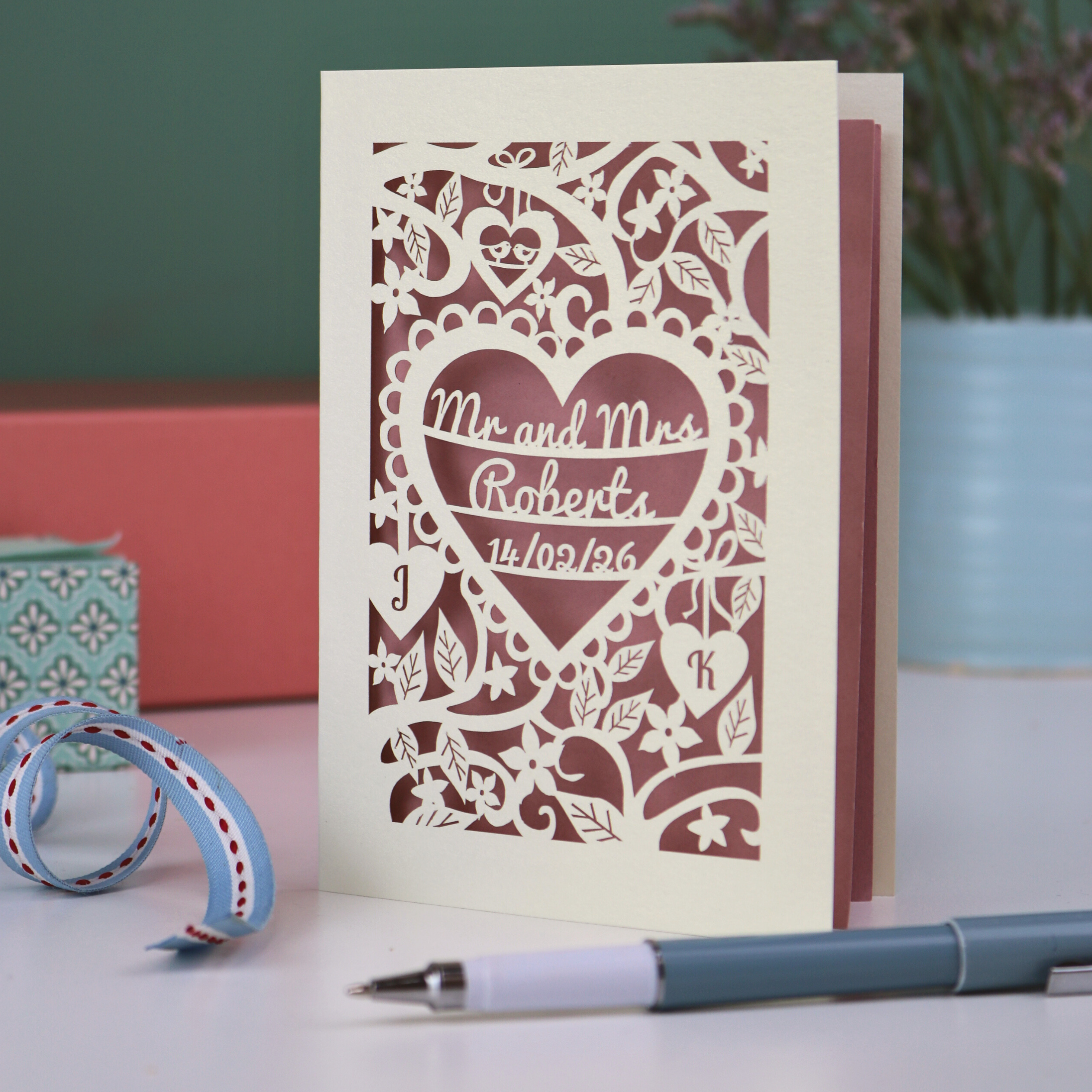 A personalised laser cut wedding card with floral heart design, sitting on a white desk with ribbons, stationery items and plant on a green background. Card is cream with "Mr and Mrs Roberts, 14/02/26" on 3 lines of text inside a heart shape with intricate floral cut out border.