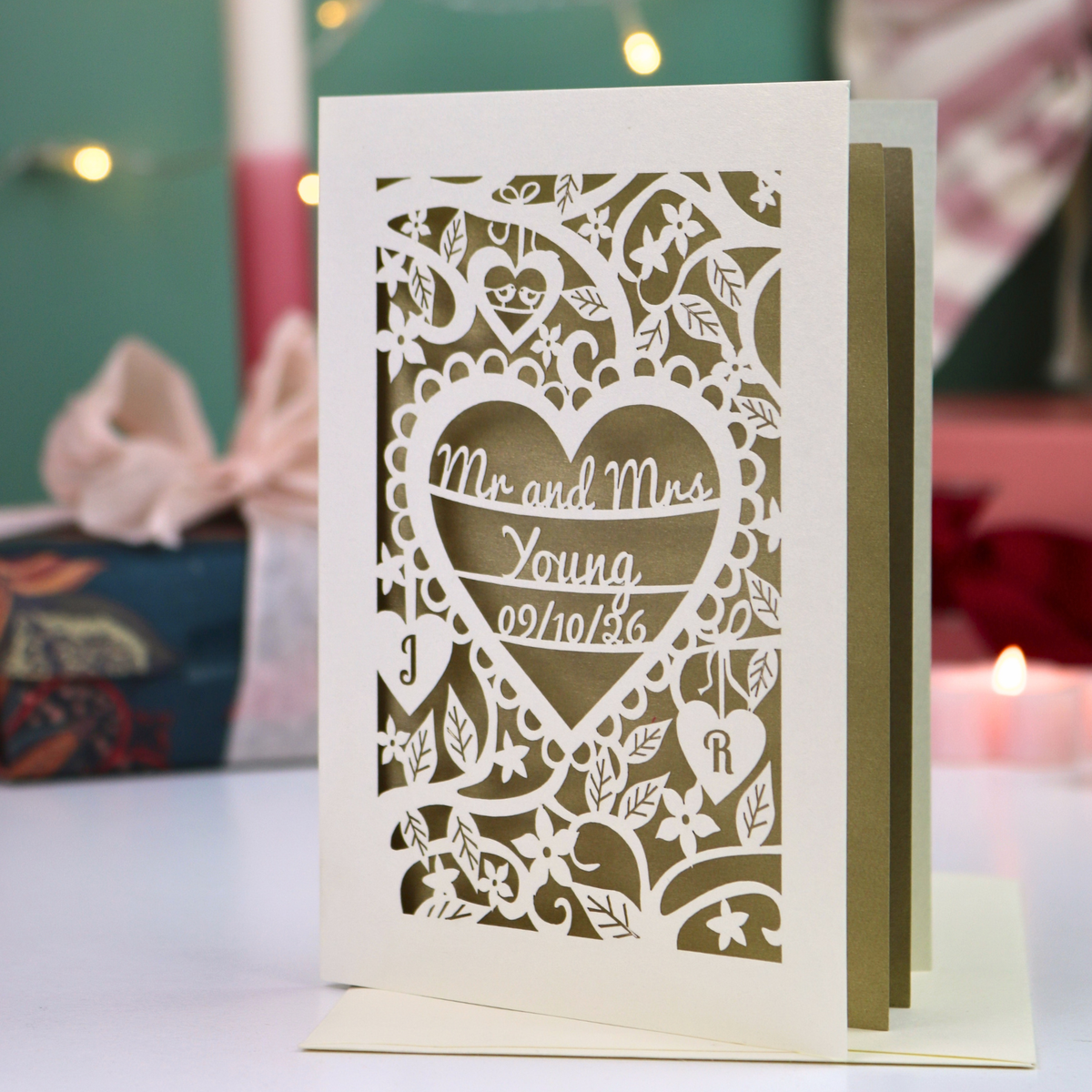 A personalized laser-cut paper card with a cream front panel featuring intricate cut out floral and heart patterns, with the text 'Mr and Mrs Young' and '09.10.26'