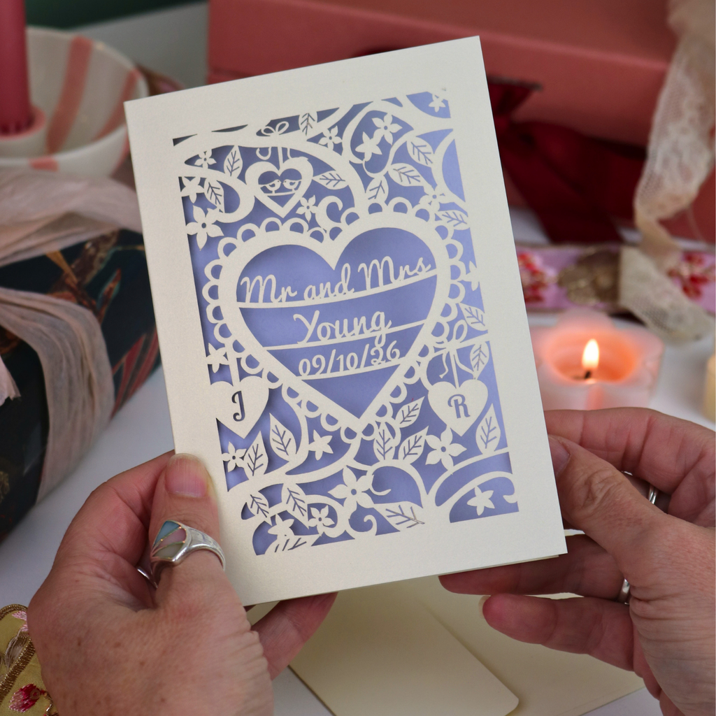 A close-up of a hand holding a personalized laser-cut wedding invitation card. The card features intricate cream cut out floral and heart patterns over a lilac background, with the text 'Mr. and Mrs. Young 09/10/26'