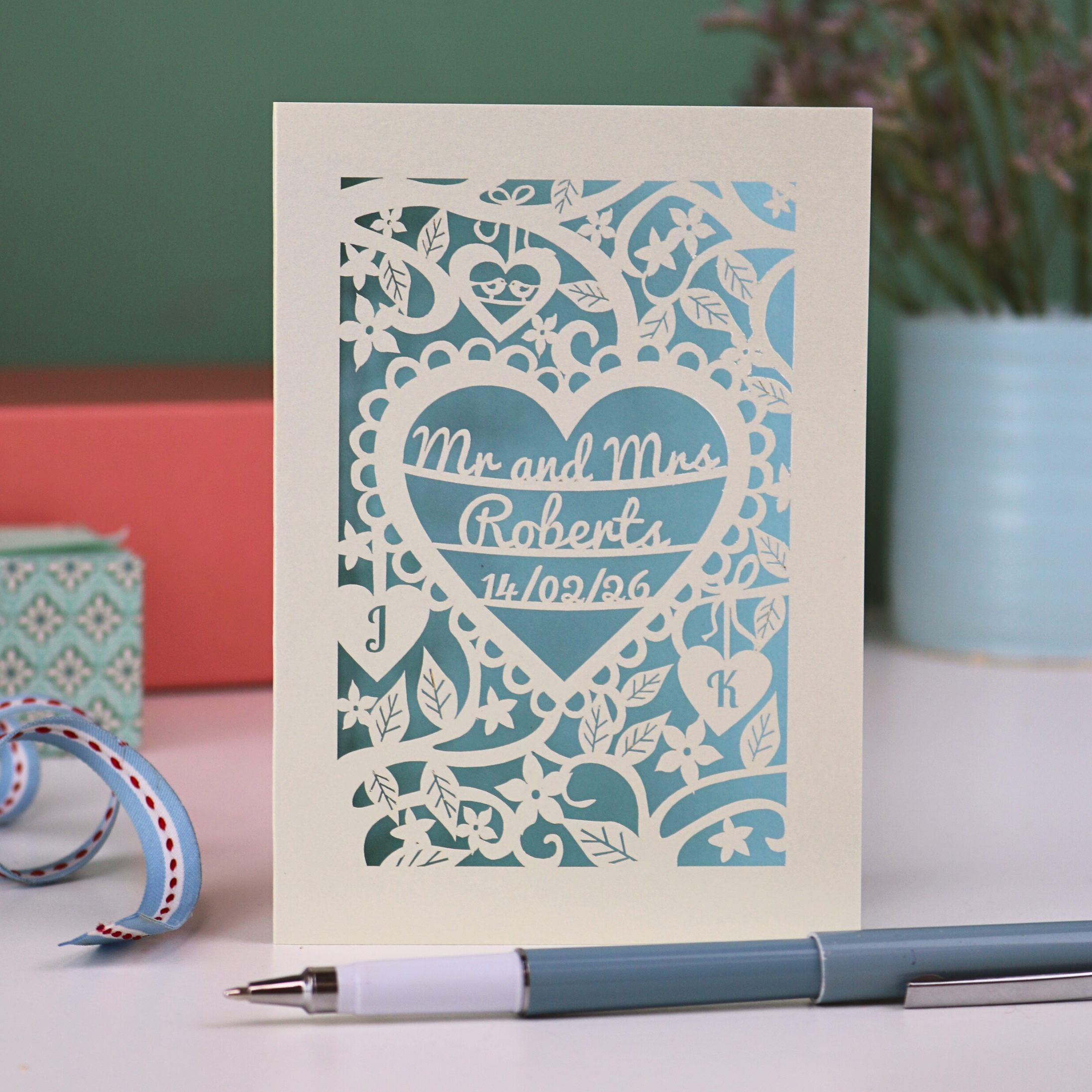 A personalised laser cut wedding card with floral heart design, sitting on a white desk with ribbons, stationery items and plant on a green background. Card is cream with "Mr and Mrs Roberts, 14/02/26" on 3 lines of text inside a heart shape with intricate floral cut out border.