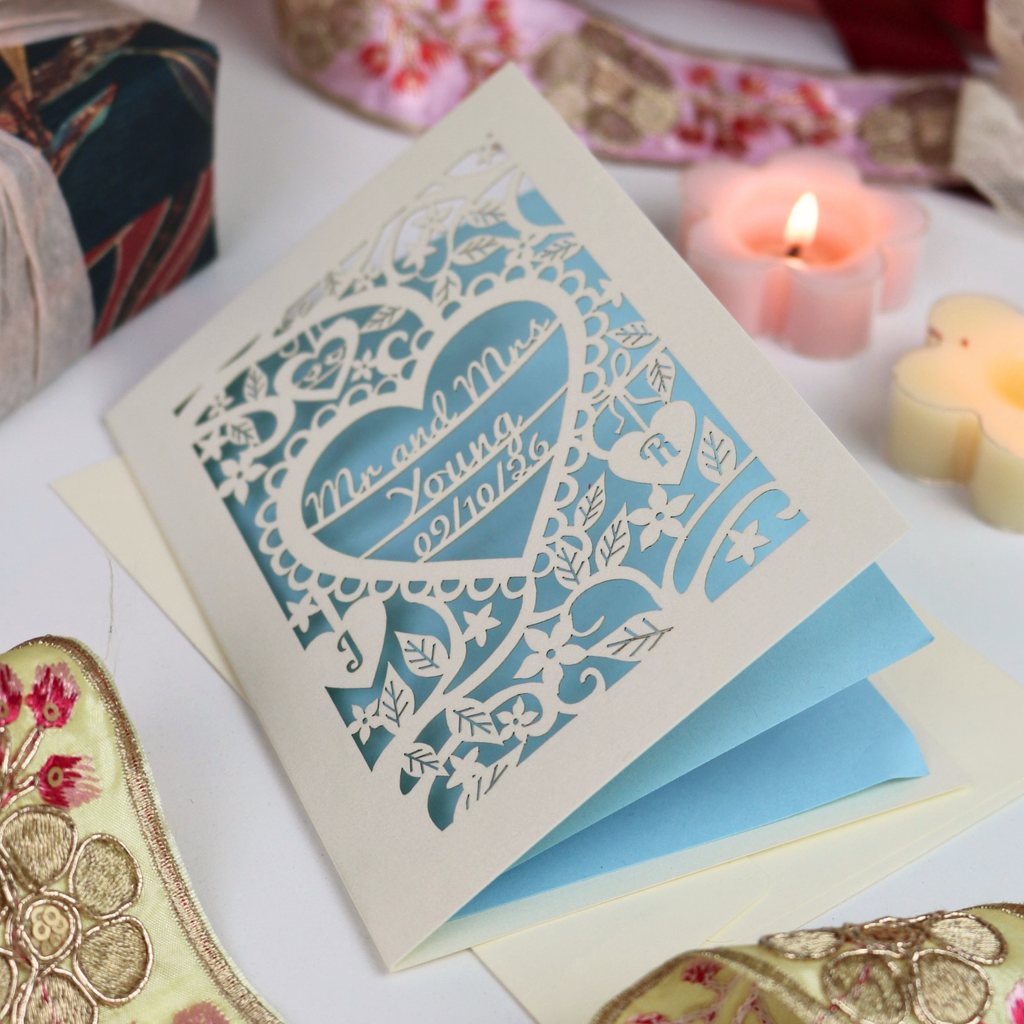 A laser-cut wedding invitation card with a heart-shaped cut out and floral design, displayed on a white surface with candles and decorative fabric in the background.