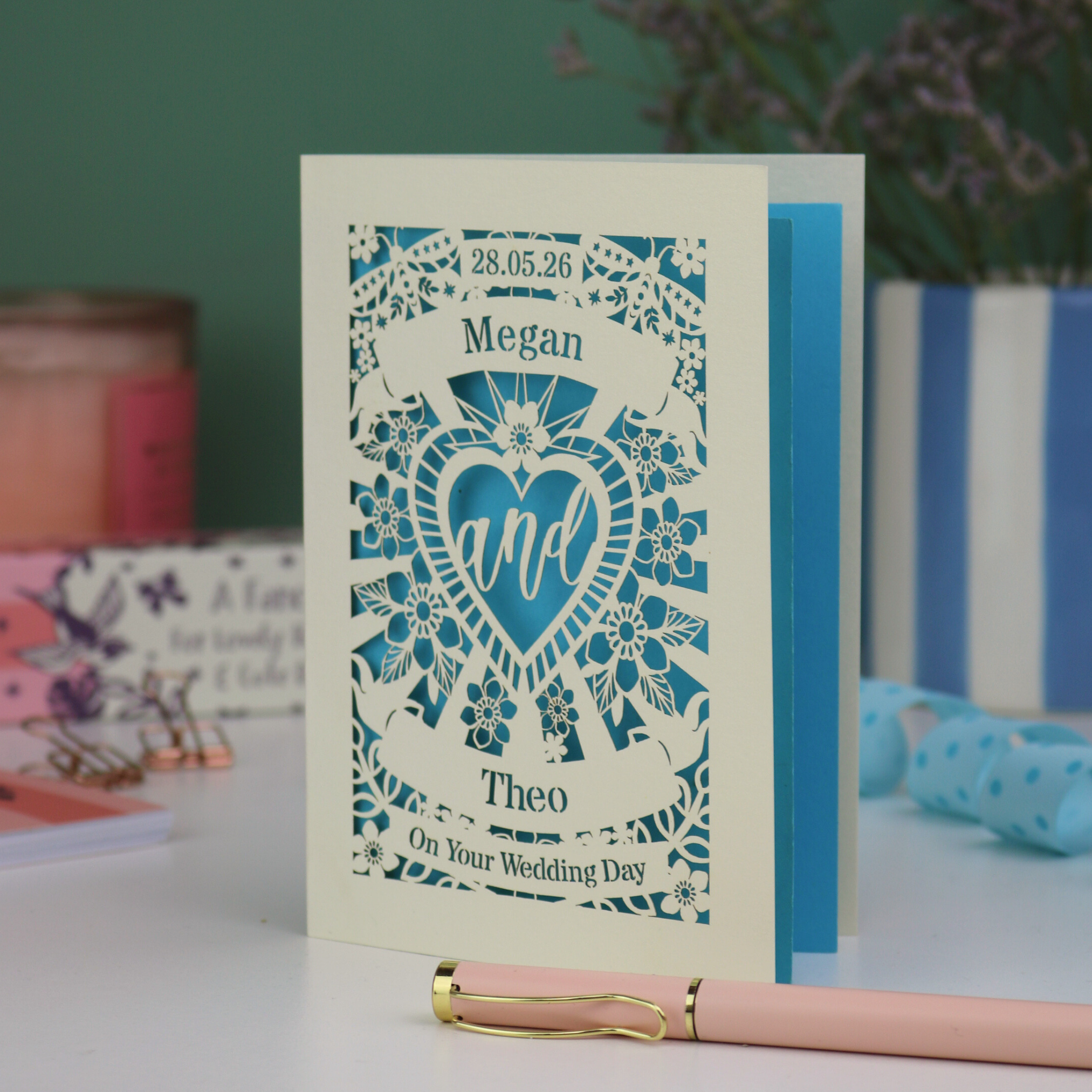 Personalised paper cut wedding card with decorative design and names on a table with candles and flowers in the background.