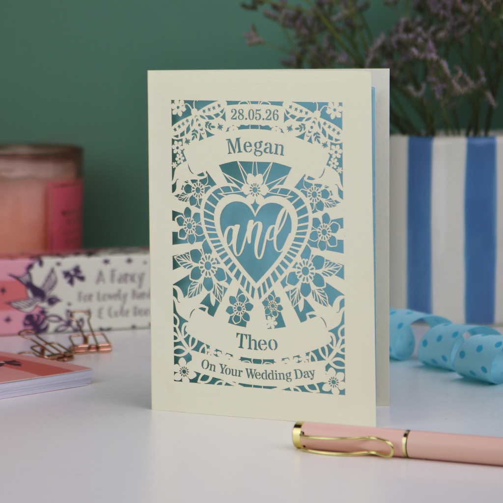 Personalised laser cut wedding card with decorative design and names on a table with candles and flowers in the background.