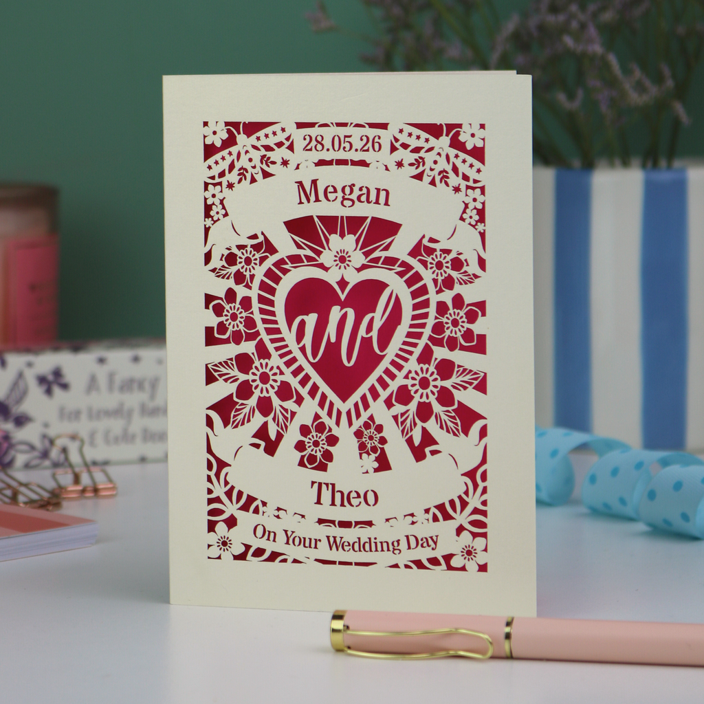 Personalised laser cut wedding card with decorative design and names on a table with candles and flowers in the background.