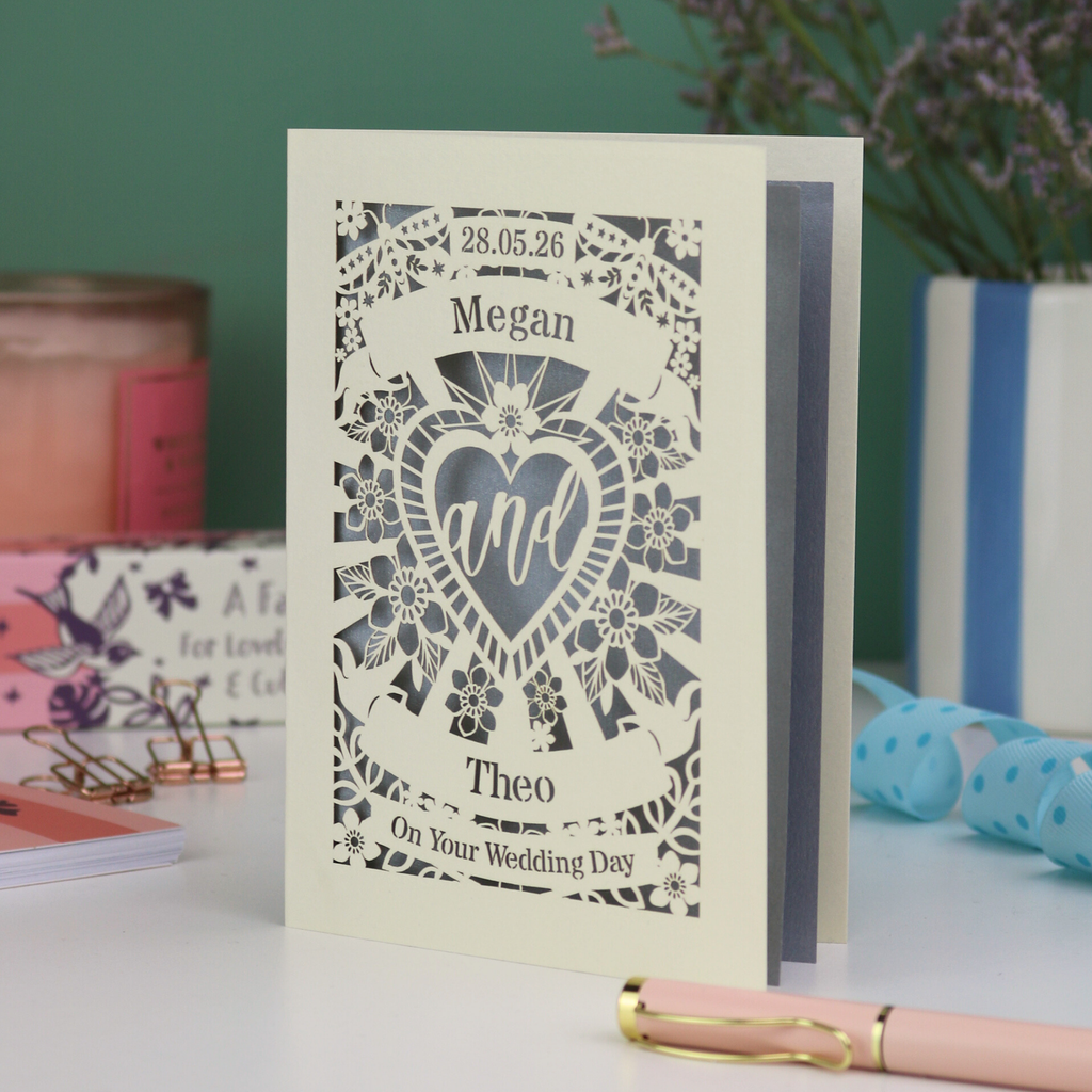 Personalised laser cut wedding card with decorative design and names on a table with candles and flowers in the background.