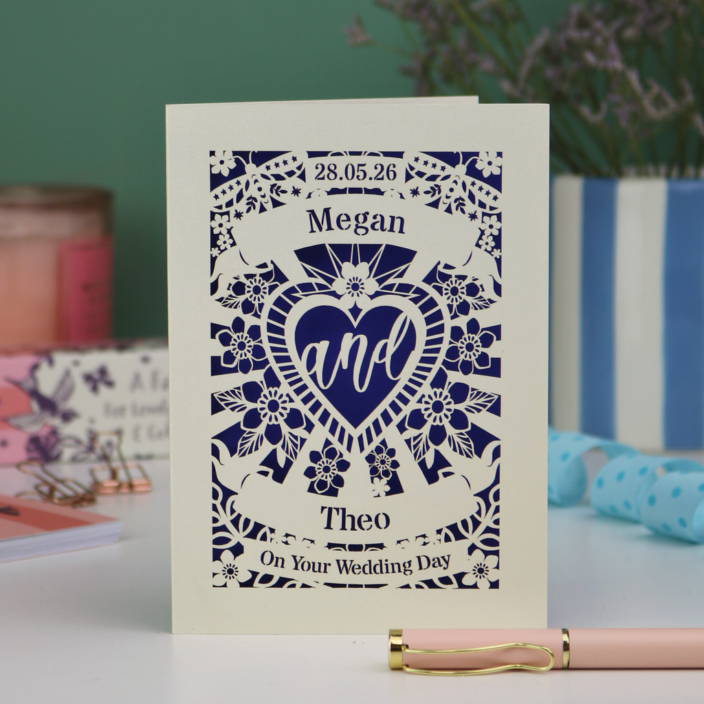 Personalised laser cut wedding card with decorative design and names on a table with candles and flowers in the background.