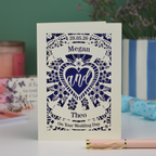 Personalised laser cut wedding card with decorative design and names on a table with candles and flowers in the background.