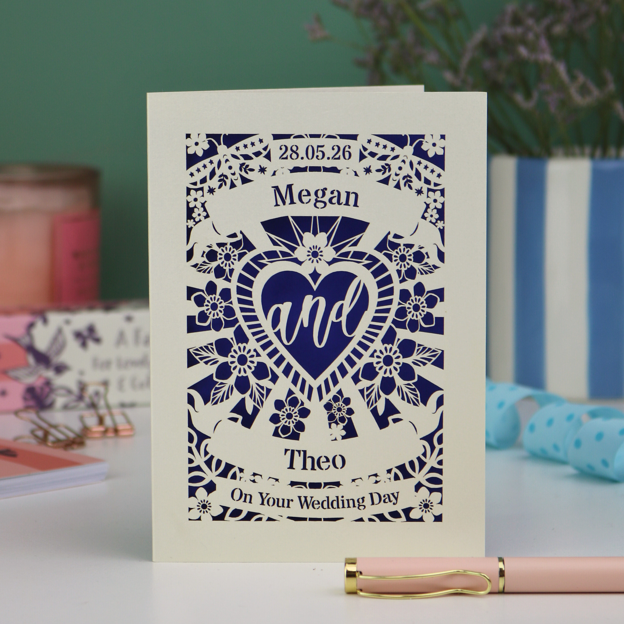 Personalised laser cut wedding card with decorative design and names on a table with candles and flowers in the background.