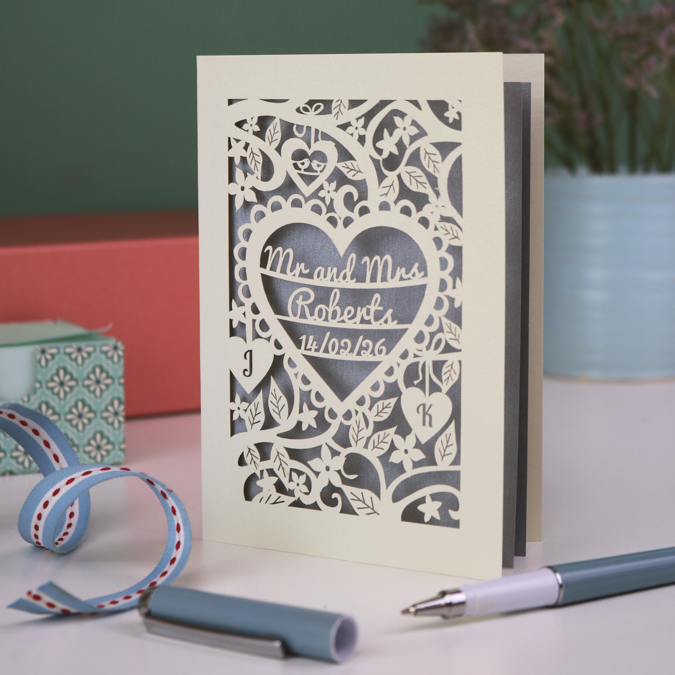 A personalised laser cut wedding card with floral heart design, sitting on a white desk with ribbons, stationery items and plant on a green background. Card is cream with "Mr and Mrs Roberts, 14/02/26" on 3 lines of text inside a heart shape with intricate floral cut out border.